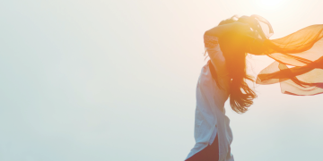 A woman in white shirt an orange scarf with arms raised behind her head walks confidently forward in the wind.